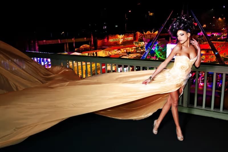 A model in a long flowing dress on a bridge at night, the funfair lights blurred behind her